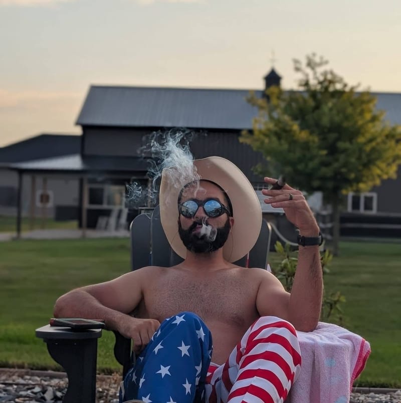 Abe reclining shirtless in a lawn chair in a straw cowboy hat and stars-and-stripes trousers, cigar smoke rising, a barn behind him.