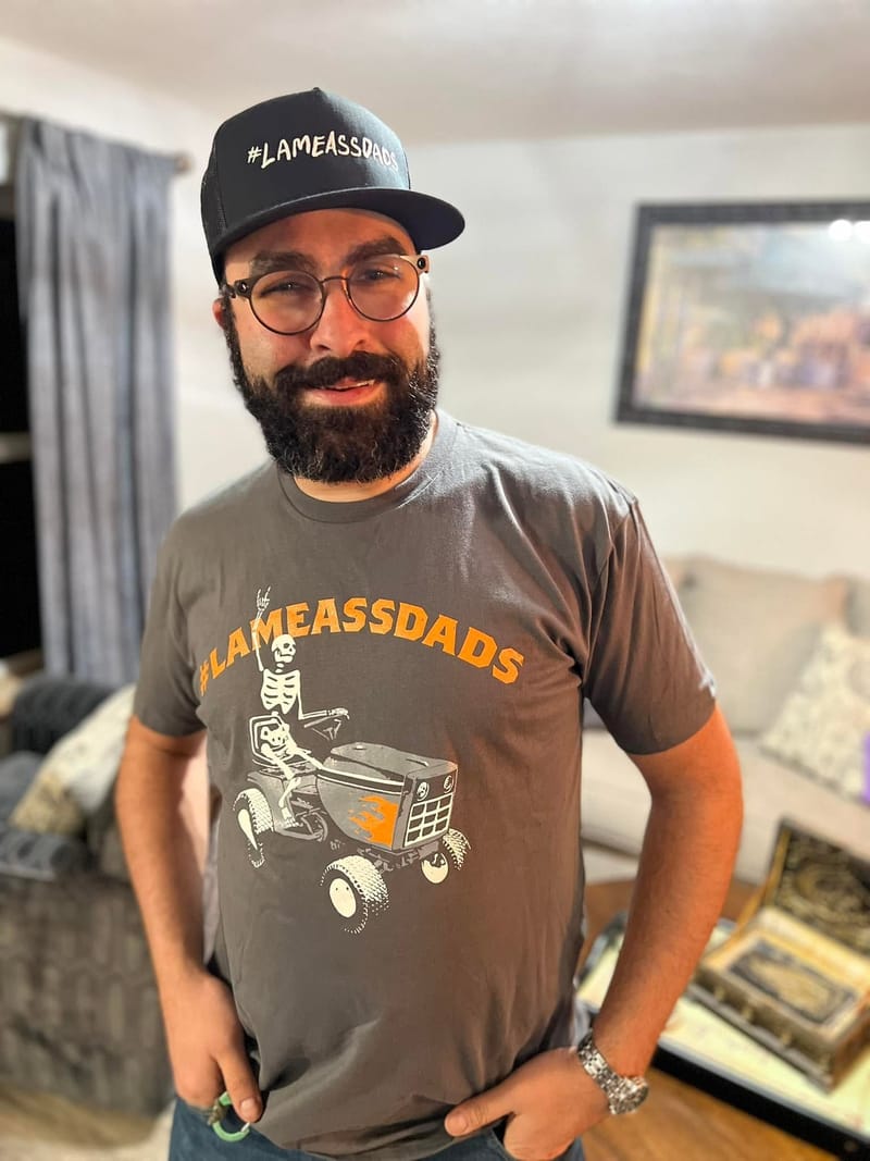 Abe indoors in a cap and a shirt reading LAMEASSDADS, hands on hips, entirely unbothered.