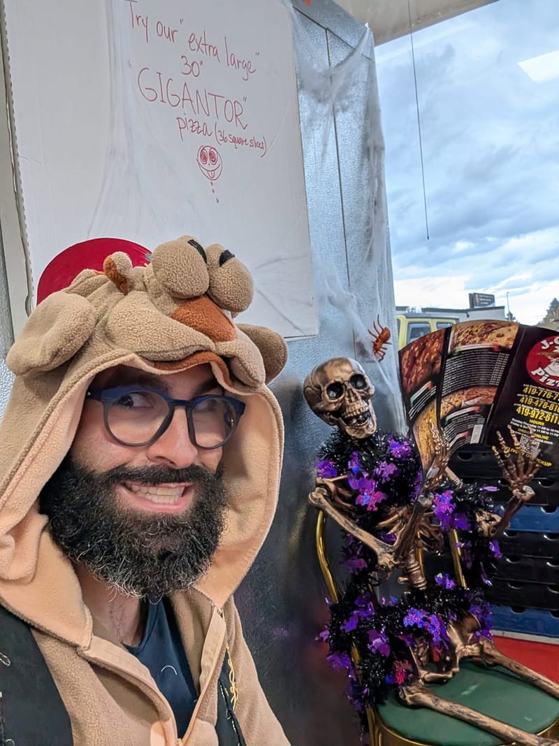 Abe in a plush animal hood beside a hand-lettered sign advertising a thirty inch Gigantor pizza, a decorative skeleton propped alongside.