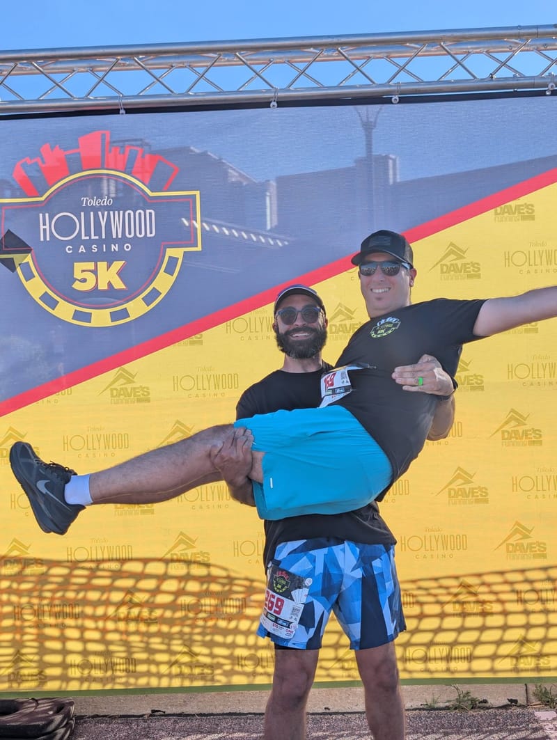 Abe being carried horizontally in another runners arms, both laughing, in front of a Toledo Hollywood Casino 5K backdrop.