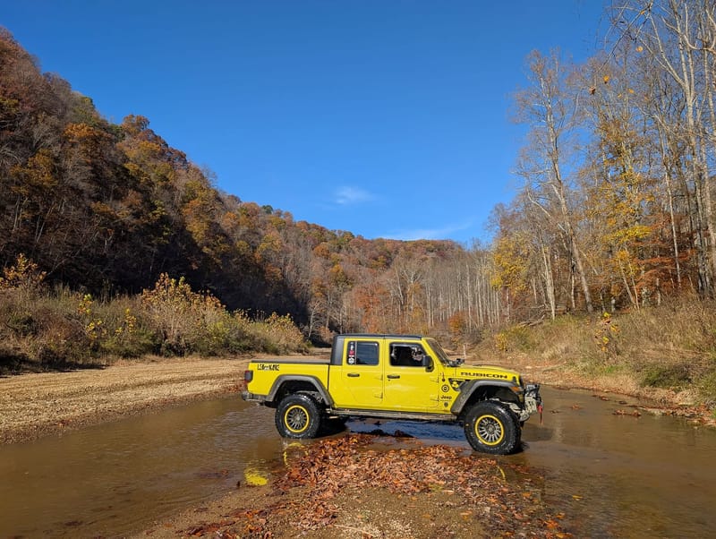 The yellow Jeep Gladiator standing in a shallow creek crossing, water round its tyres, autumn hillside rising behind.