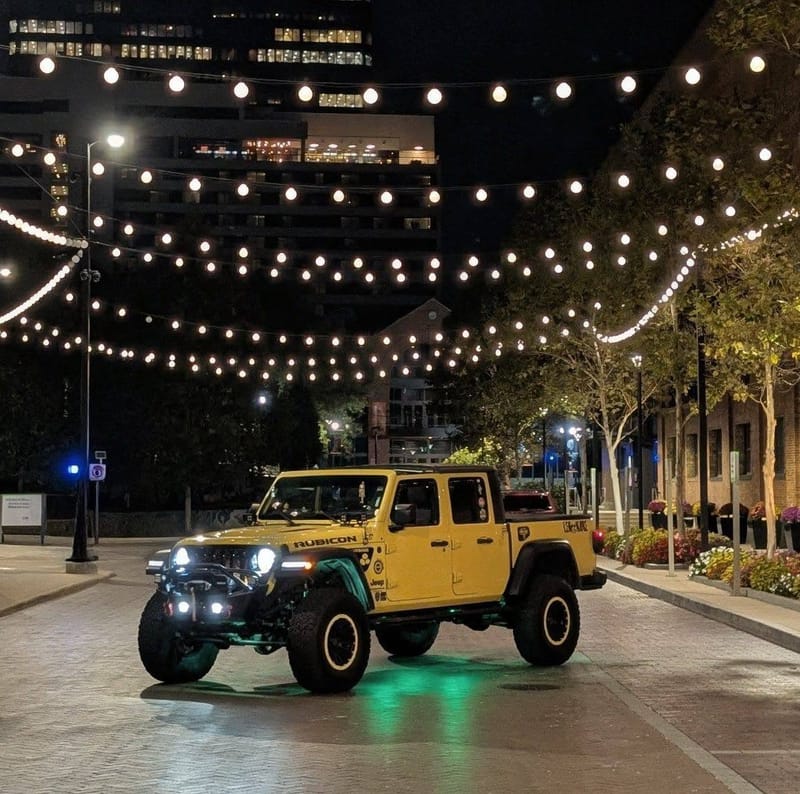 The yellow Jeep at night on a downtown street under strings of cafe lights, green underglow lit.