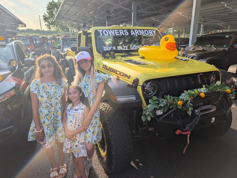 The yellow Jeep at an outdoor show with a rubber duck on the hood and his daughters standing in front of it.