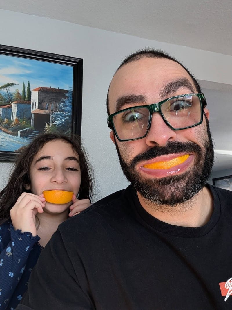 Abe and one of his daughters side by side, each grinning around an orange slice held in their teeth.