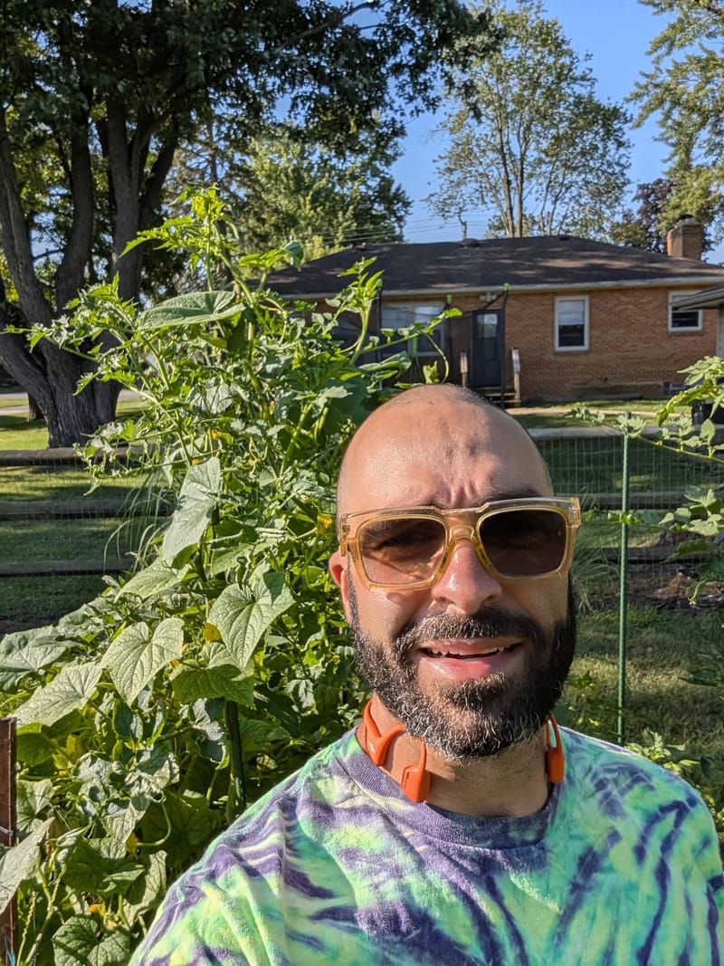 Abe in sunglasses and a tie-dye shirt standing among tall leafy garden plants in a back yard.