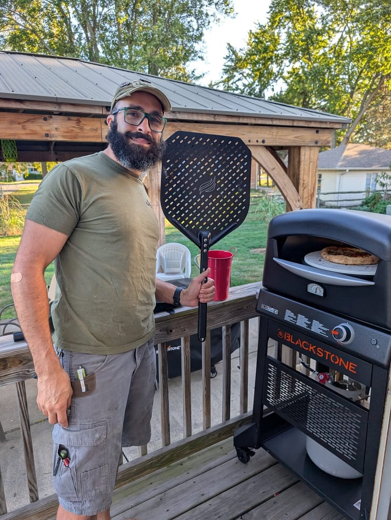 Abe on a back deck beside a Blackstone flat-top griddle, drink in one hand, griddle press in the other.