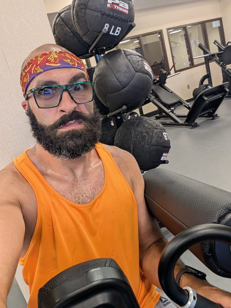 Gym selfie in an orange tank top with a rack of weighted medicine balls behind him.